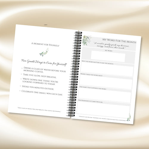 A spiral-bound journal or planner shown open on a soft beige satin-like background. The left page has motivational text and small watercolor botanical illustrations, and the right page has a monthly word reflection section with blank lines and headings.