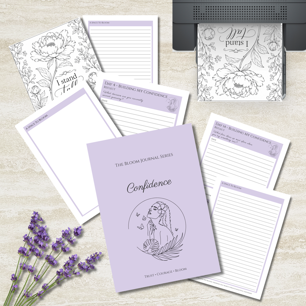 A flat-lay of a journaling and affirmation kit featuring a lavender hardcover journal titled 'Confidence' and several matching lined notepads and printable pages with floral line art and lavender accents, arranged on a light wood surface with lavender sprigs and a printer visible in the background.