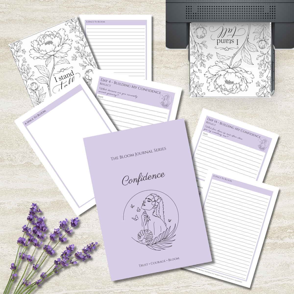 A flat-lay of a journaling and affirmation kit featuring a lavender hardcover journal titled 'Confidence' and several matching lined notepads and printable pages with floral line art and lavender accents, arranged on a light wood surface with lavender sprigs and a printer visible in the background.
